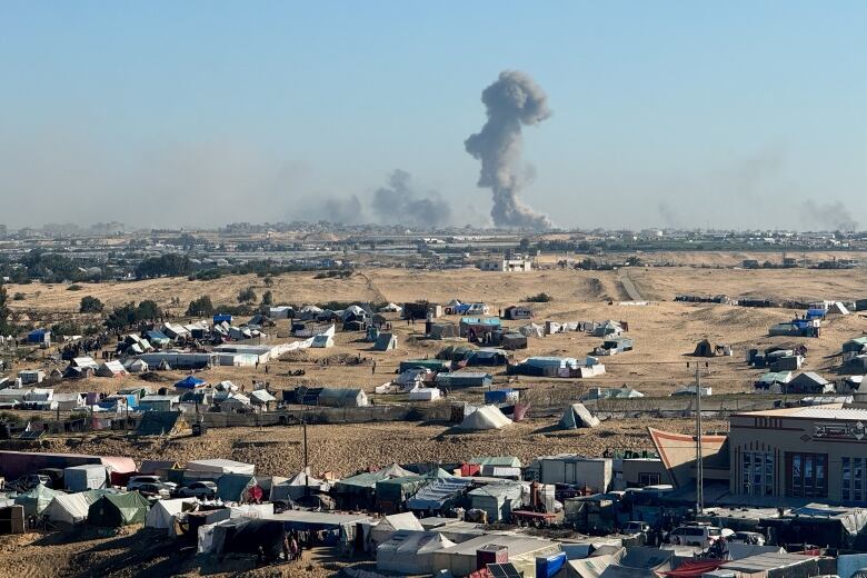 Smokes rises from a city in the background as tents are seen in the foreground.