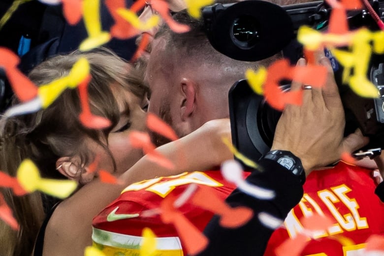 Amid orange and yellow tickertape, a cameraman records as a woman with her eyes closed and a man wearing a football sweater that says KELCE on the back kiss.