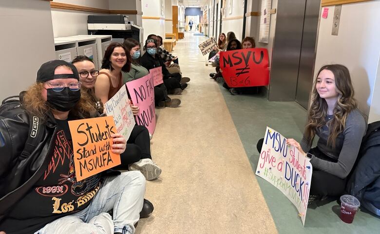 Students sit on the ground in a hallway holding signs.