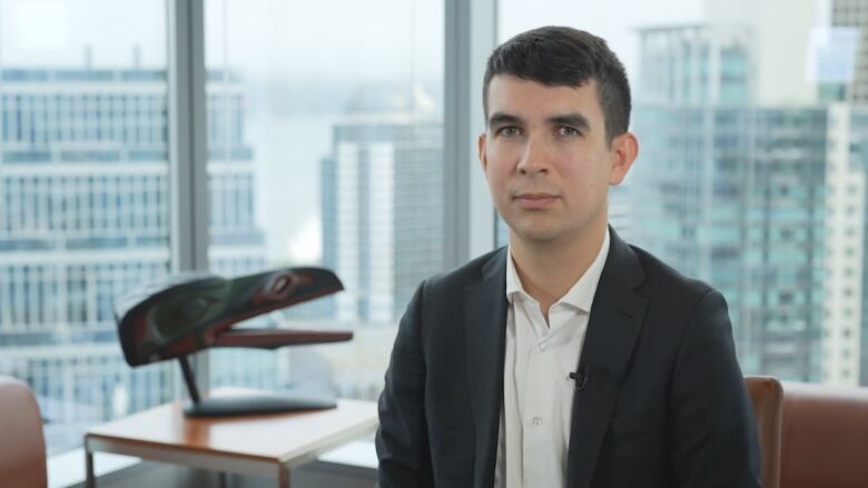 A man with short brown hair looks into the camera seated in a confrence room. He wears a dark blue wuit jacket and white coloard shirt. 