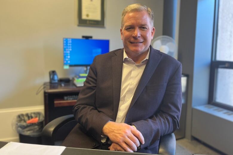 Man with white hair in business jacket and open-necked shirt, sitting in an office, smiling into the camera.