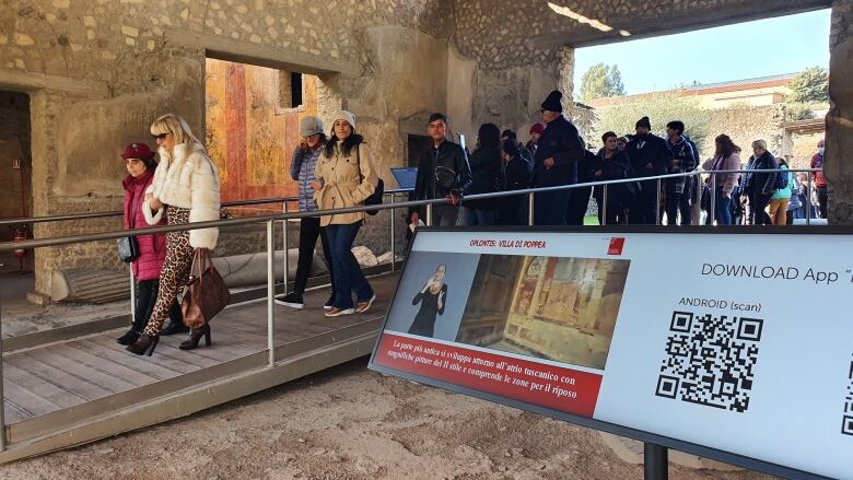 People walking through an archaeological museum pass by a monitor that includes a sign-language interpreter as well as a QR code and some text in Italian.