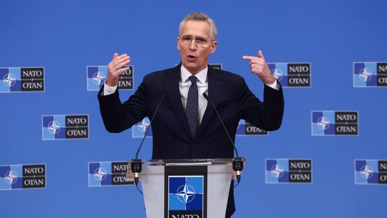 A clean-shaven, bespectacled man in a suit and tie gestures with arms wide apart while speaking at a podium. 