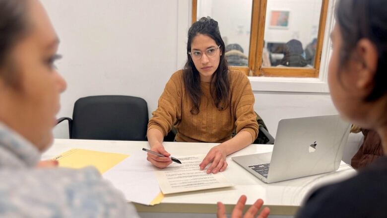 A community worker sits at a desk with a sheet of paper, facing two clients with their backs to the camera.