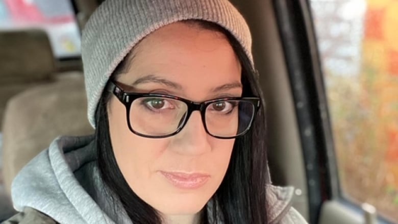 A woman in a grey toque and black-framed glasses sits inside a vehicle.