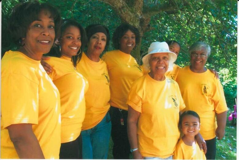 A group of 7 women and a young child are all wearing bright yellow shirts and smiling at a camera.