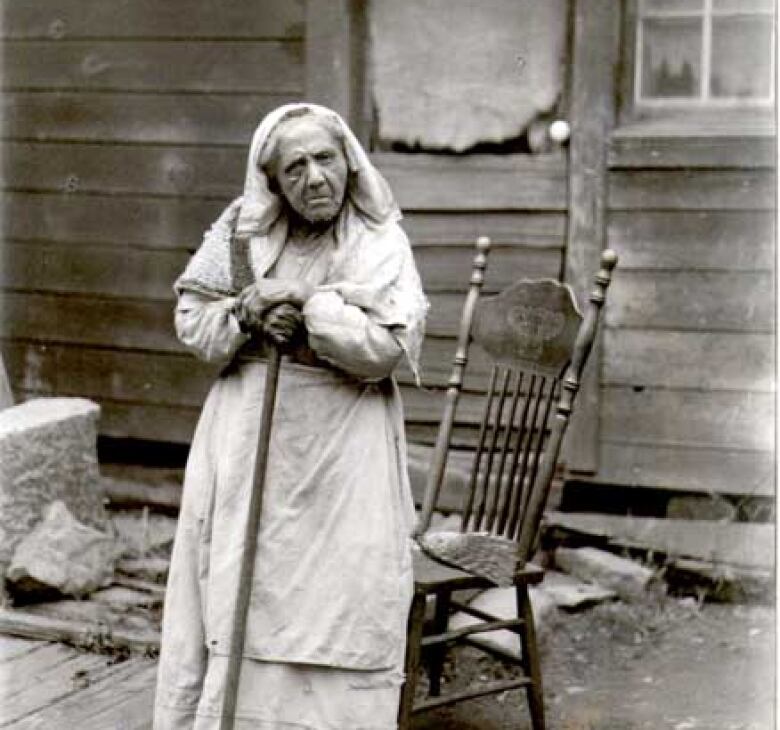 In a black and white photo an elderly woman is standing and holding a cane in front of a wooden chair and a wooden house. 