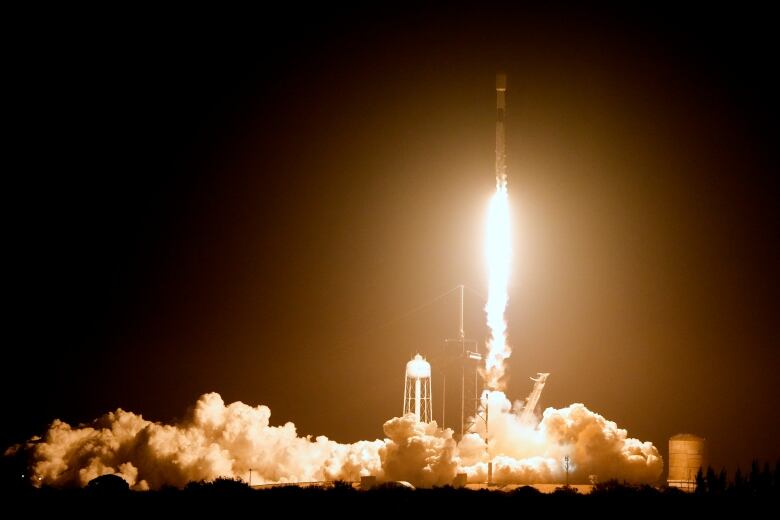 A dark sky is lit up by large volumes of smoke and a rocket ship leaving a platform.