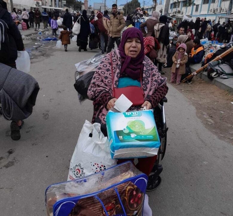 An old woman sits in a crowded street, surrounded by her belongings in bags.