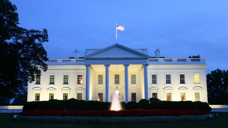 An exterior view of the White House lit up in the early evening with a U.S. flag waving in the wind on the roof and a fountain and flower bed in the foreground. 