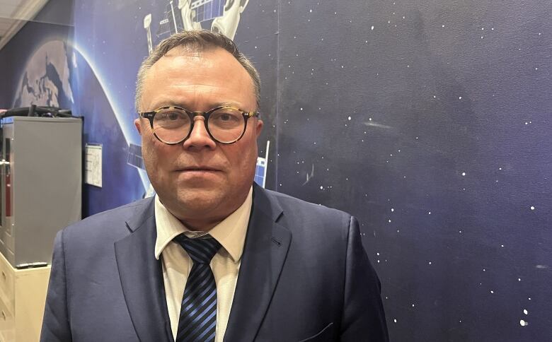 A man with short, gray hair wearing a blue three-piece suit stands for a photo. He has round glasses, a round face and a solemn expression.