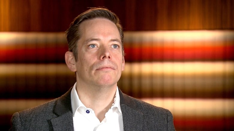 A man in a business suit is pictured with an orange, yellow and red wall in the background.