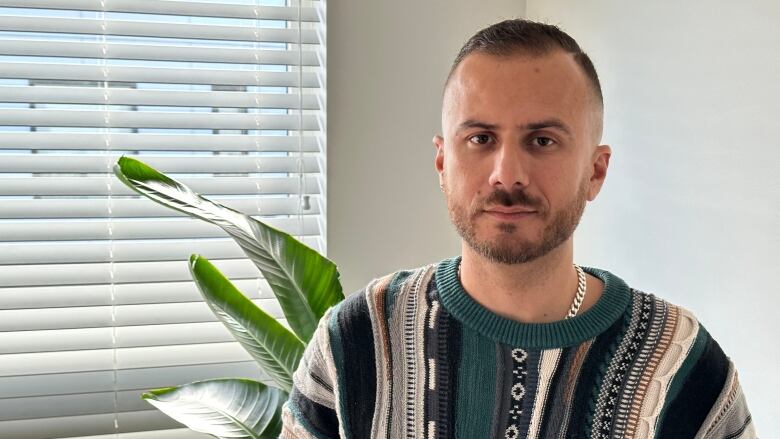 A man in a stripe-patterned knit sweater looks unsmilingly at the camera while standing next to a window indoors, with leaves from a large green plant seen behind him.