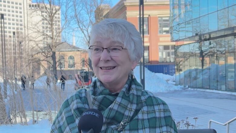 a woman wearing a tartan shawl speak into a microphone on the sidewalk in winter. 