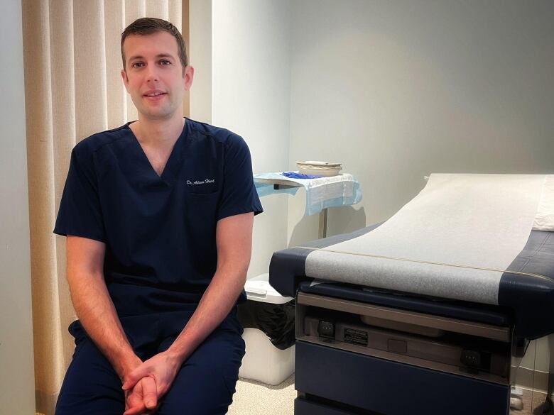 Young man in dark blue scrubs sits in medical room.