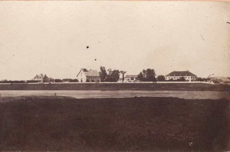 Black and white photo, looking across a river, shows five buildings on the opposite bank.