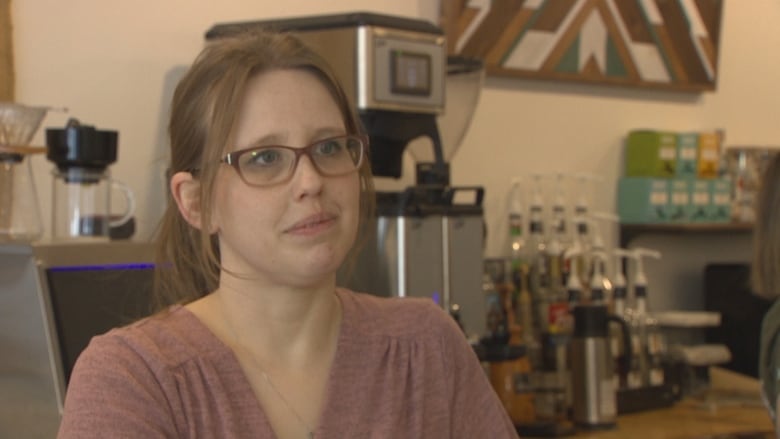 A white woman with light brown hair is seen inside a cafe. she is wearing a pink t shirt and glasses.