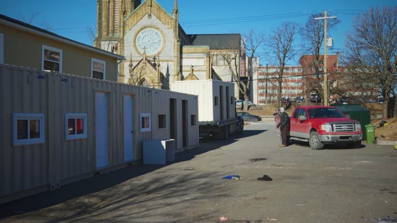 A shipping container structure is loaded into a parking lot under supervision by a man in a red truck. 