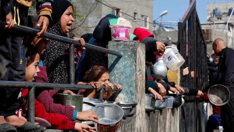 Children waiting for food, with bowls and buckets in their hands, standing behind metal rails along a concrete wall. 