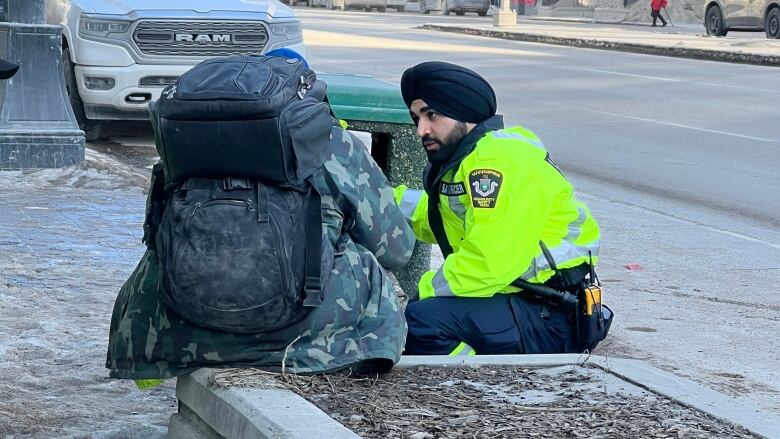 A man wearing a turban is crouching down on a sidewalk. He's talking to a man sitting with his back to the camera.