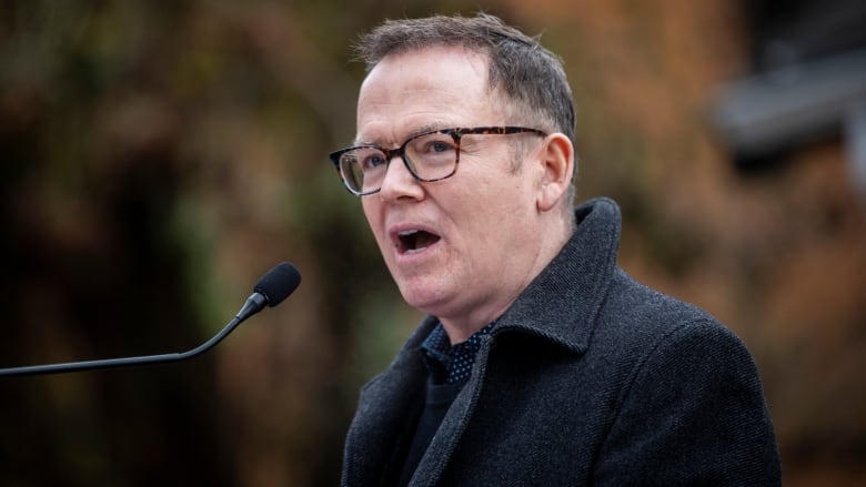 A white man with black hair, wearing glasses, speaks in an outdoor environment.