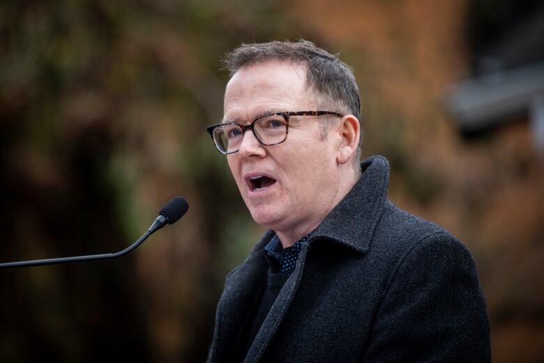 A white man with black hair, wearing glasses, speaks in an outdoor environment.