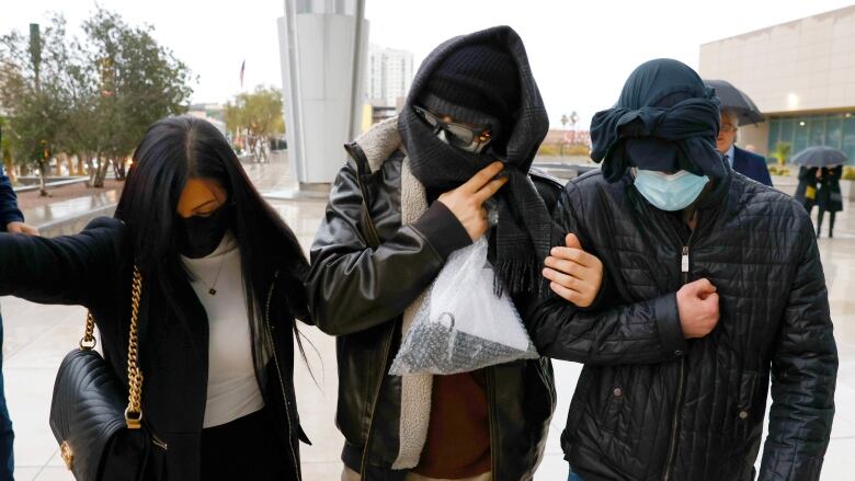 A man covers his face with a hood and mask and dark sunglasses while walking between a man and a woman on a sidewalk.