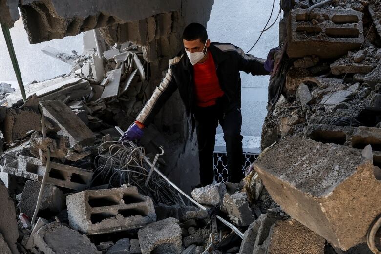 A man wearing a mask appears to tread carefully inside a dwelling that has been heavily damaged, with large concrete blocks and other debris shown.