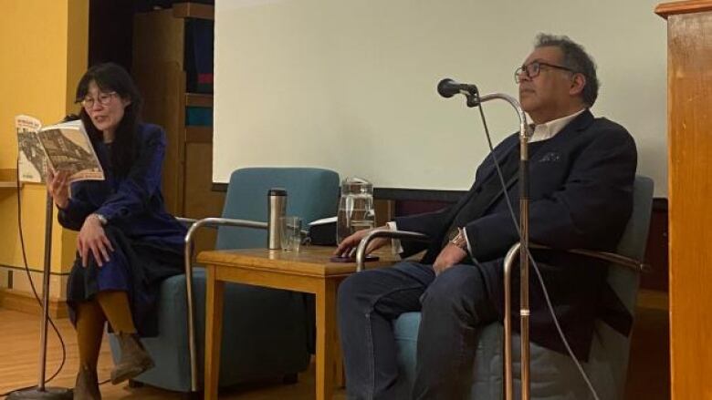 An Asian woman with long dark hair reads from a book as she sits in a chair onstage across from a seated Brown man with grey hair and glasses.