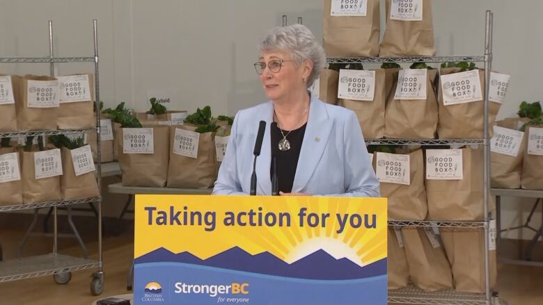 A woman in a suit with grey hair and glasses stands in front of shelves with grocery bags on them.