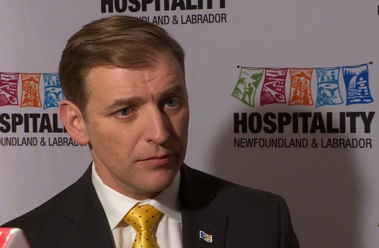 A man wearing a suit and a yellow tie stands in front of a white screen with logos reading, 