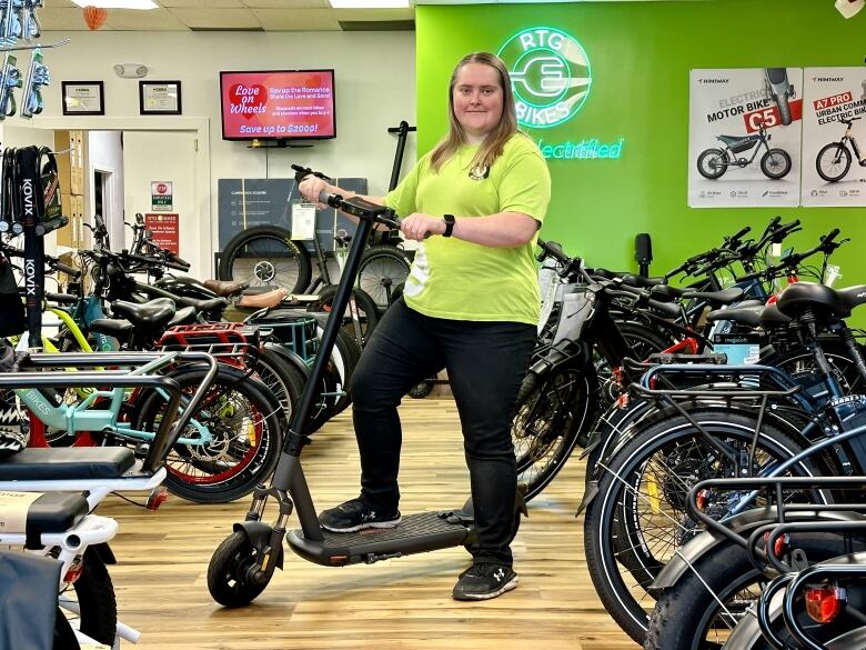 A woman wearing a bright green t-shirt stands on an electric kick scooter in a store filled with bicycles.