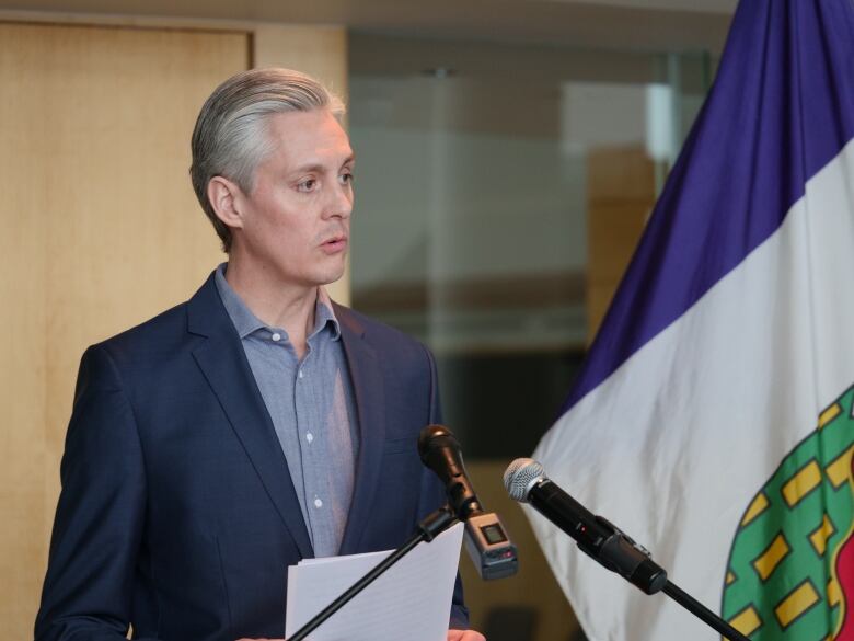 A man in a suit stands before 2 microphones, speaking.