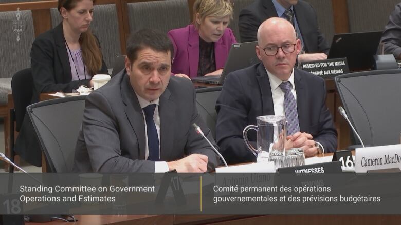 Two men in suits sit in a committee room on Parliament Hill answering questions from MPs. 