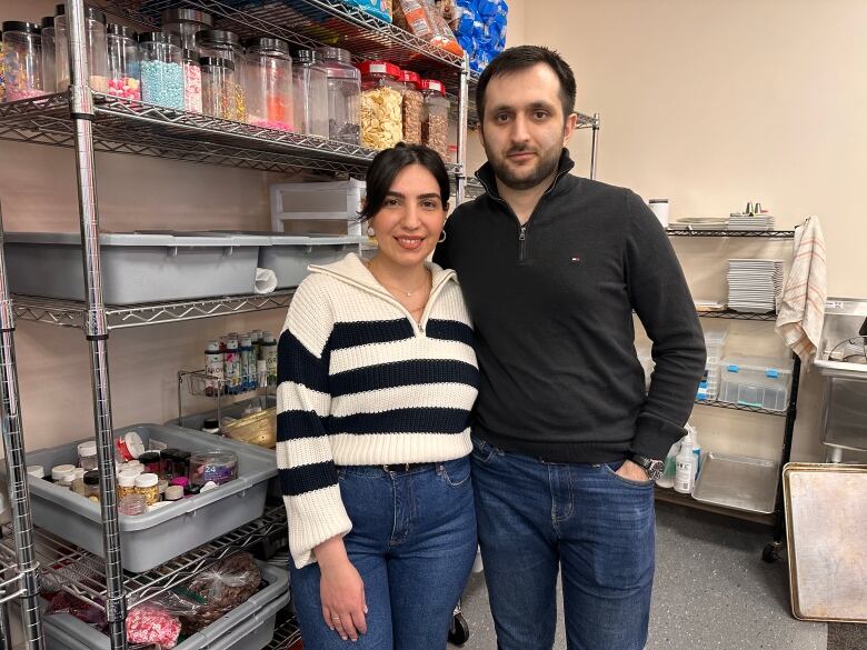 A woman in a black and white striped sweater and a man in a black long sleeve shirt stand in front of metal cabinets full of baking supplies. 