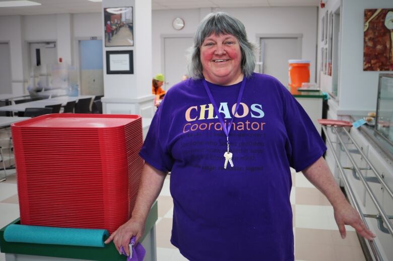 A smiling woman in a purple t-shirt stands behind a stack of red cafeteria trays. 