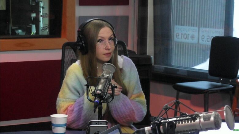 Woman in a rainbow coloured sweater wearing round glasses sits in front of a microphone.