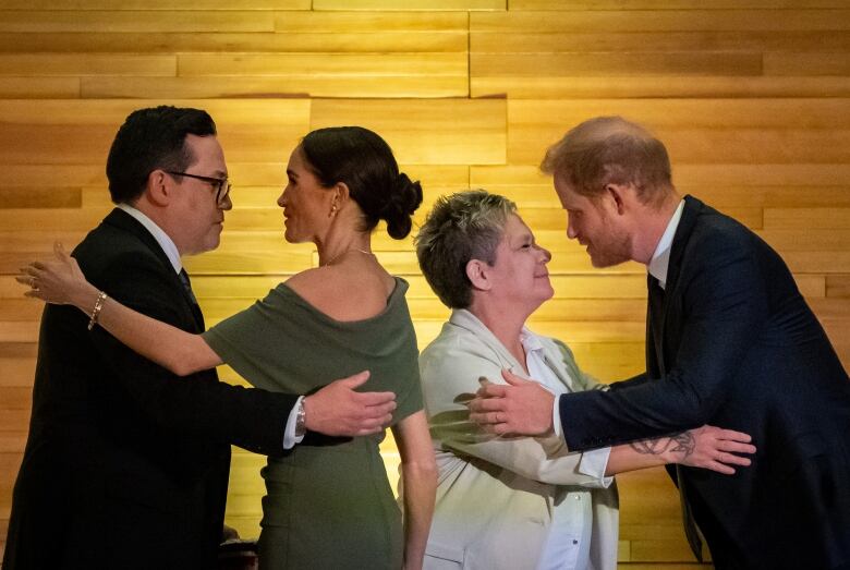 Two people greet two other people in front of a wood wall.