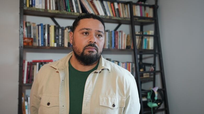 A man in a beige button down and green t-shirt seen looking off to the right, stands before a metal bookcase full of books. 