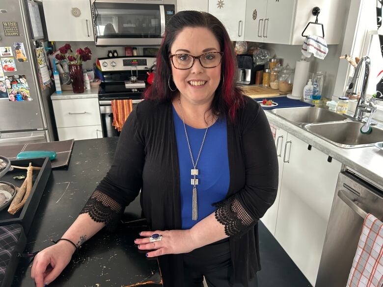 A woman with long dark hair and a blue shirt stands in her kitchen. 