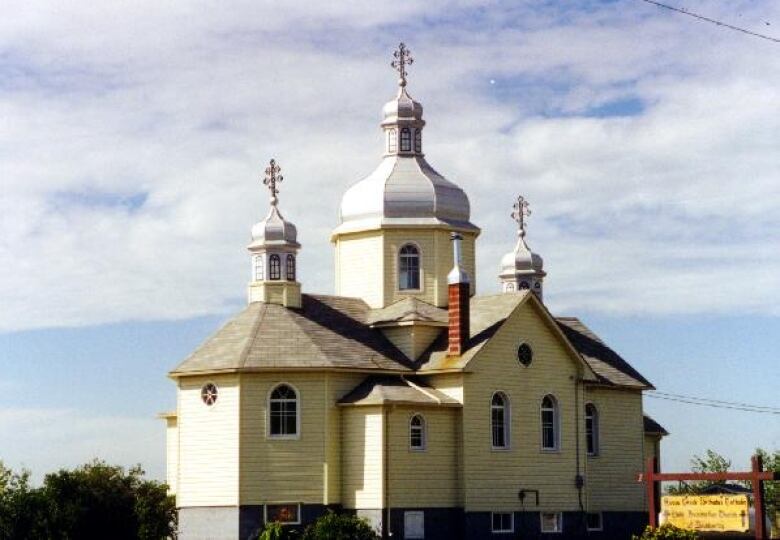 A yellow-ish church with three bright white domes is seen against the backdrop of a blue sky with many white wispy clouds. 