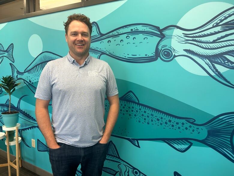 A young man in a pale blue t-shirt stands in front of a mural of squid and fish.