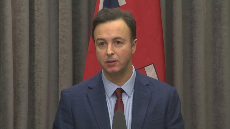 A man in a blue suit, light blue shirt and red tie sits behind a desk where a microphone is placed.