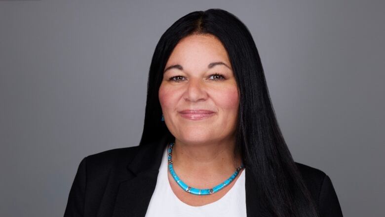 An Indigenous woman with long, straight black hair is smiling with her lips closed. A turquoise necklace wraps around her neck. She is wearing a white top under a black blazer. She is standing in front of a grey background.