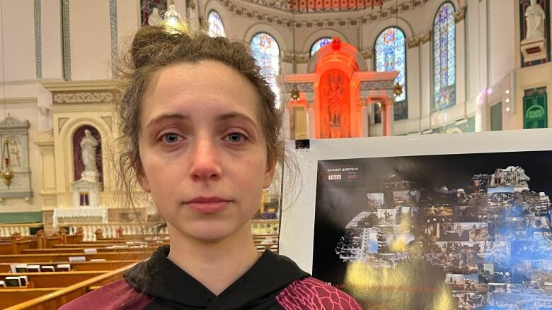 A woman stands inside a church, in front of a poster with the outline of Ukraine, filled with photos of destruction from the country.