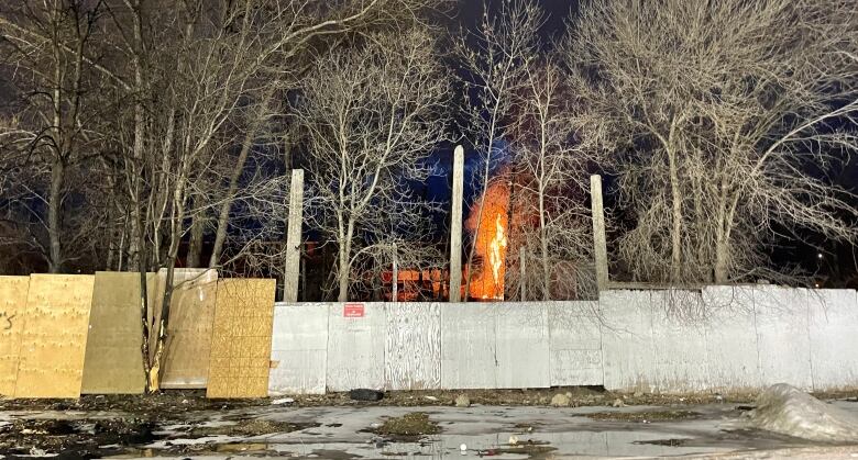 A fire can be seen burning behind a plywood fence.