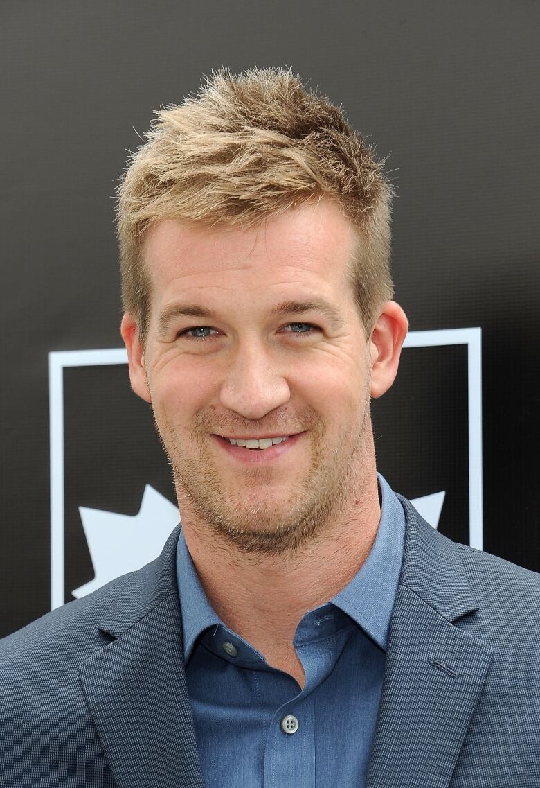 A man with stubble wearing a blazer and collared shirt is shown in closeup.