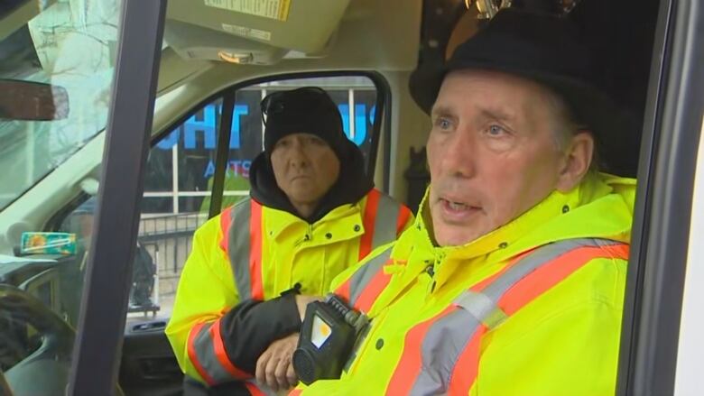 Two men wearing bright, reflective jackets sit inside the cab of a large van.