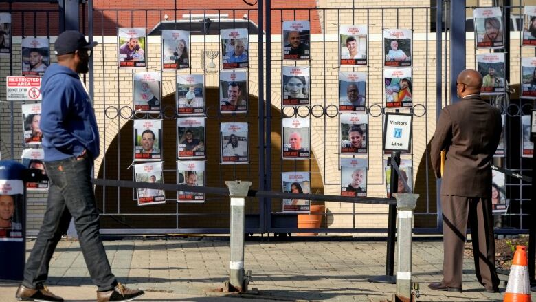 Two men stand in front of iron gates with posters of people on them