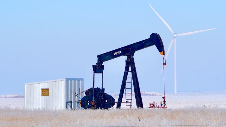 A wind turbine is pictured in the distance behind an oil pumpjack in a field.